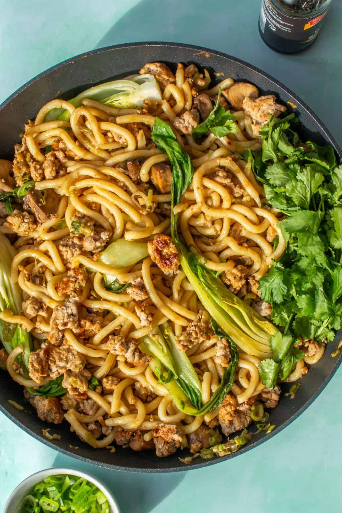 Udon noodles in a large pan with browned mince, pok choi,sliced spring onion, a lime wedge and coriander on a place blude background with a small bowl of spring onions in partial view.