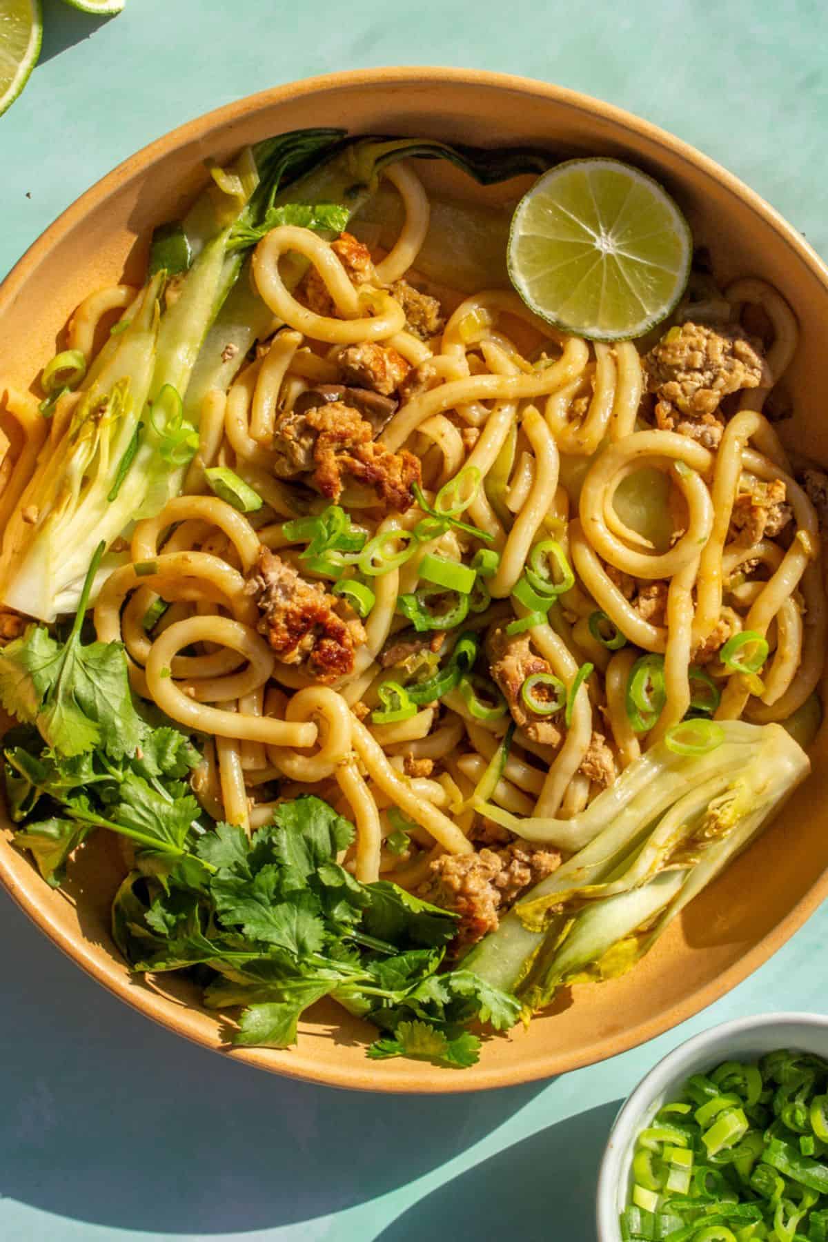 Udon noodles in a bowl with browned mince, pok choi topped withsliced spring onion, a lime wedge and coriander on apale blue background with a small bowl of spring onions in partial view.