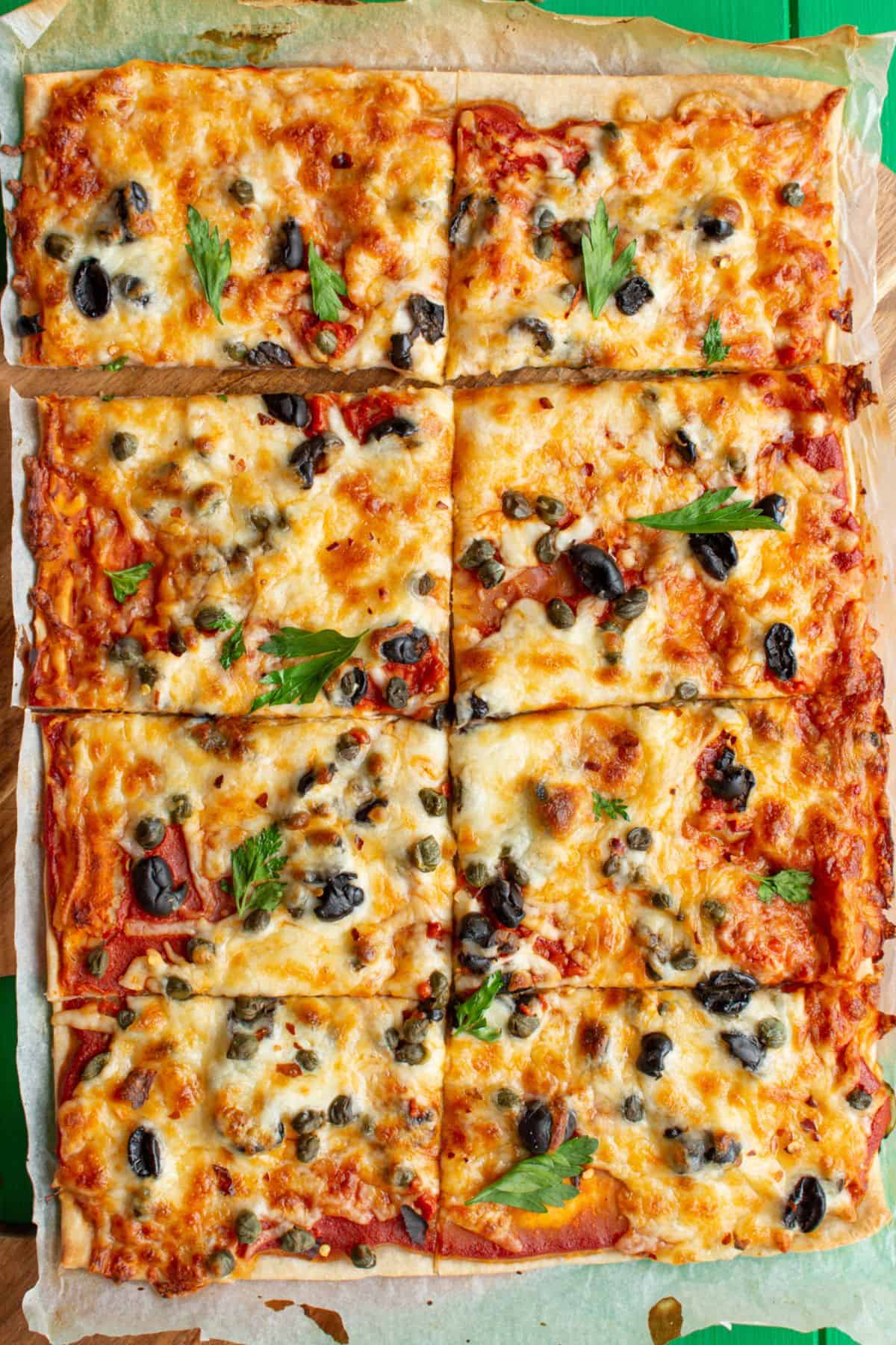 A rectangular sheet of pizza with cheesy topping, with capers and black olives and fresh parsley on parchment paper.