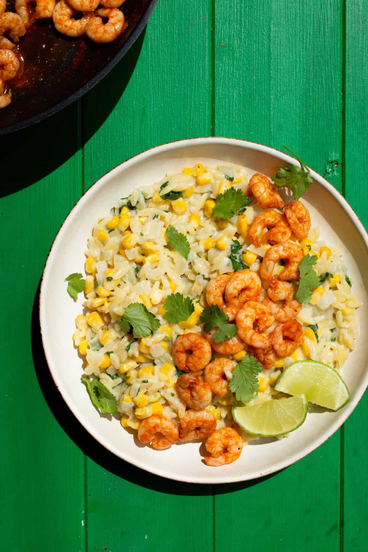 A bowl with orzo, sweet corn, spicy prawns topped with lime wedges and coriander with the pan in partial view behind on a green background.