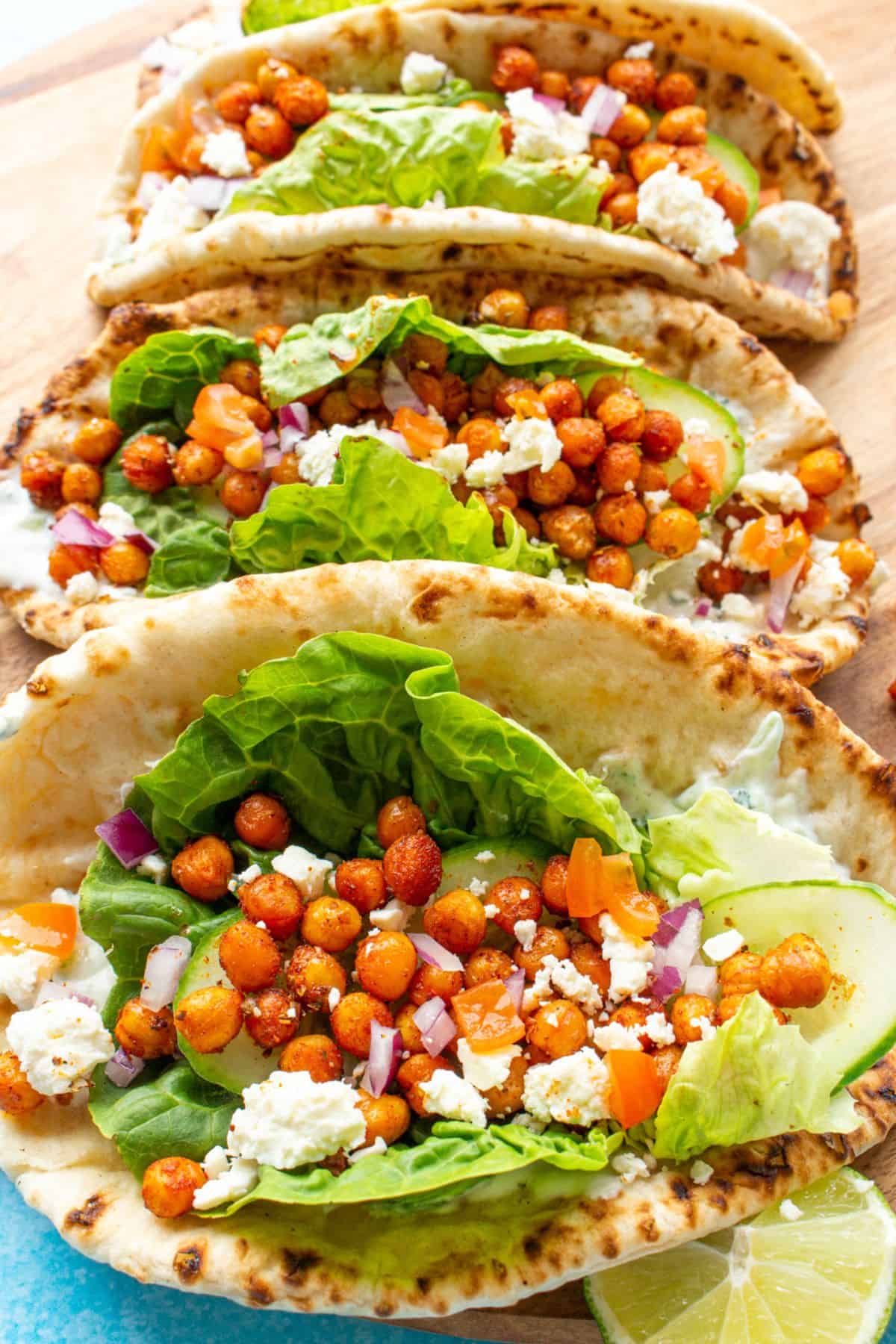 Overhead shot of 4 flat breads on a wooden board filled with roasted chickpeas, feta, red onion, lettuce and yogurt.