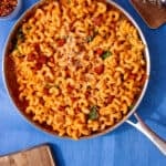 Spirali pasta in tomatoey sauce with pieces of chocken and chorizo in a large stainless steel pan on a blue background next to a chopping board with chorizo bits, a bowl with chilli and some grated cheese on a little board next to the pan.