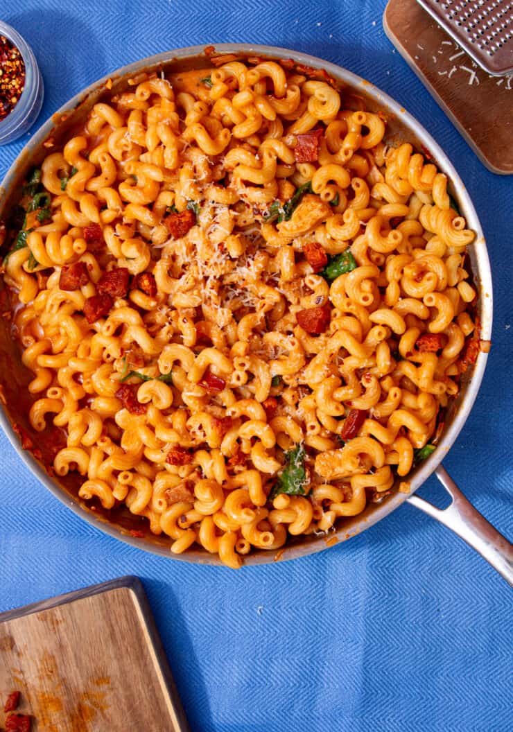 Spirali pasta in tomatoey sauce with pieces of chocken and chorizo in a large stainless steel pan on a blue background next to a chopping board with chorizo bits, a bowl with chilli and some grated cheese on a little board next to the pan.