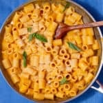 A large pan with rigatoni pasta with a butternut squash sauce and wooden spoon on a blue background.
