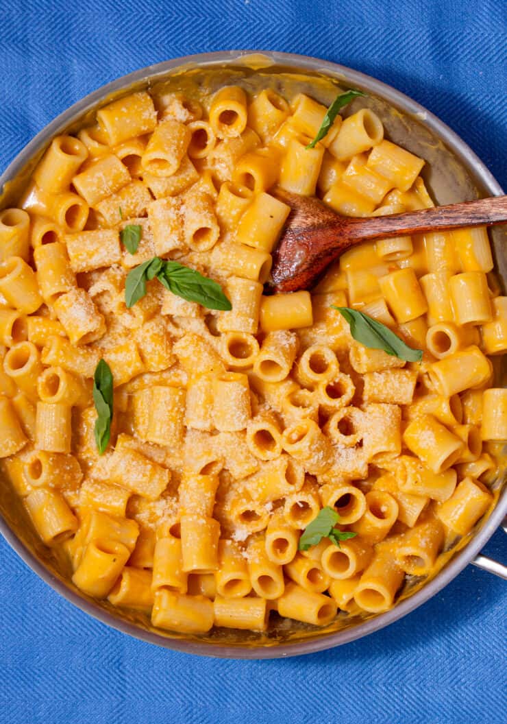 A large pan with rigatoni pasta with a butternut squash sauce and wooden spoon on a blue background.