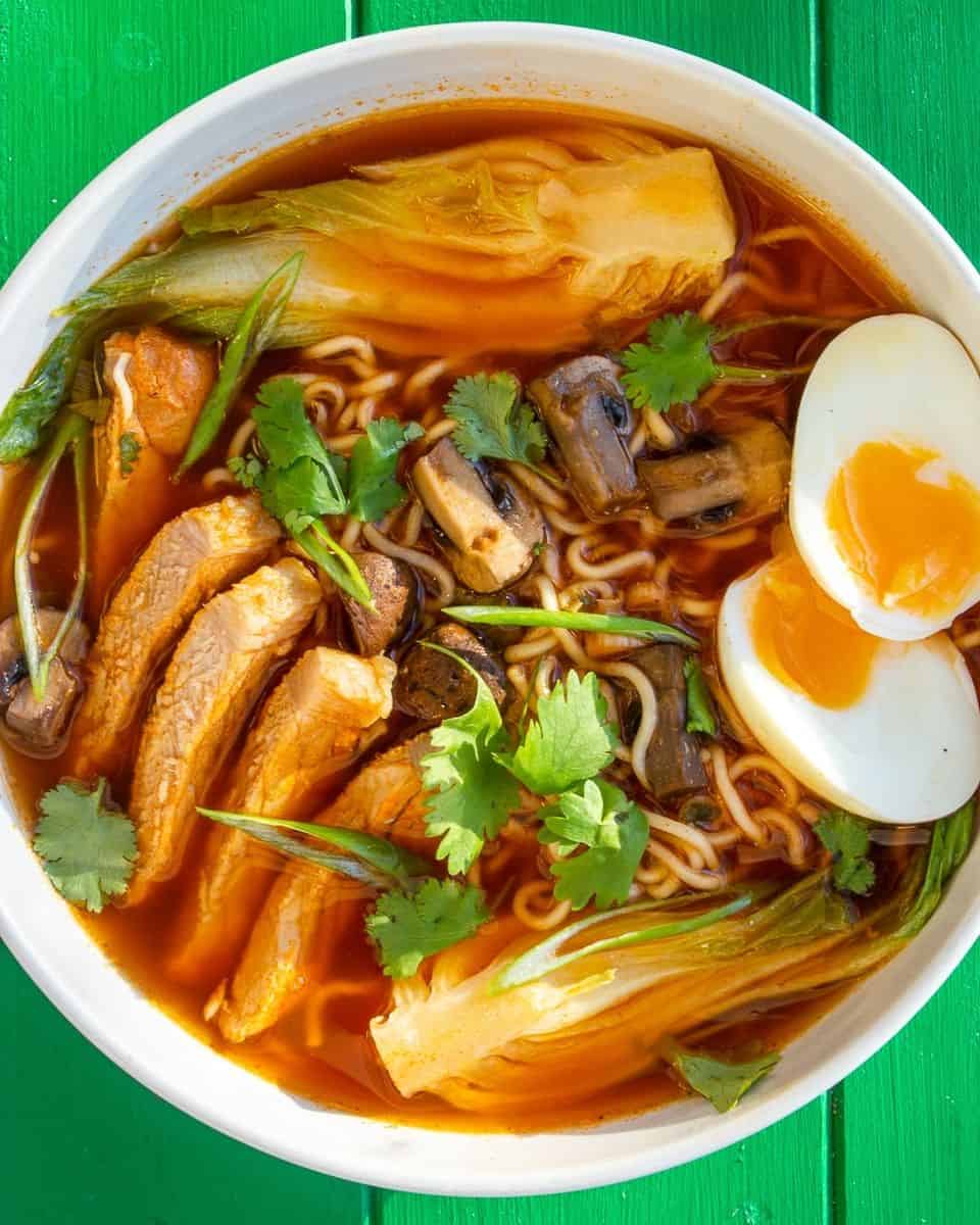 A white bowl of ramen with sliced pork, boiled egg sliced in half and coriander to top