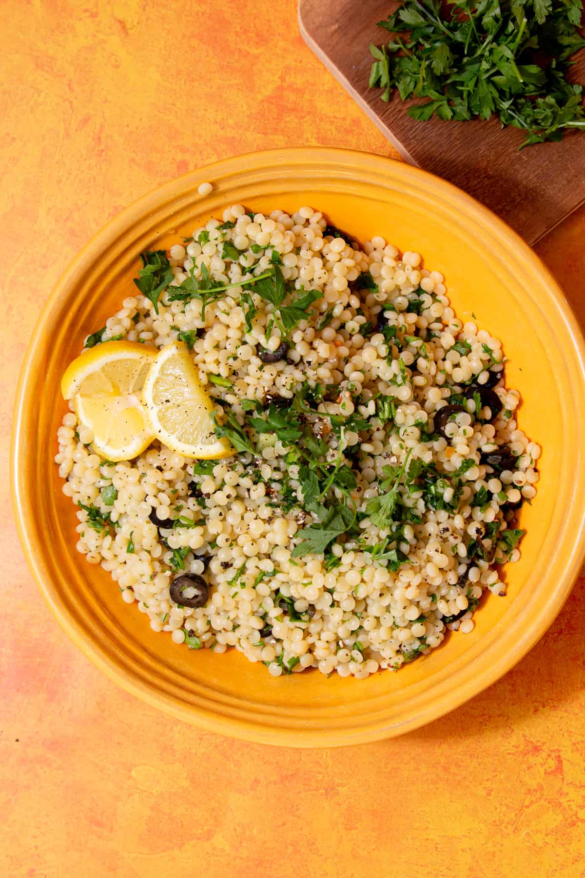 Overhead shot of a yellow dish with giant cous cous topped with herbs and lemon wedges and slices of black olives on an orange background with some fresh parsley on a chopping board in partial view.