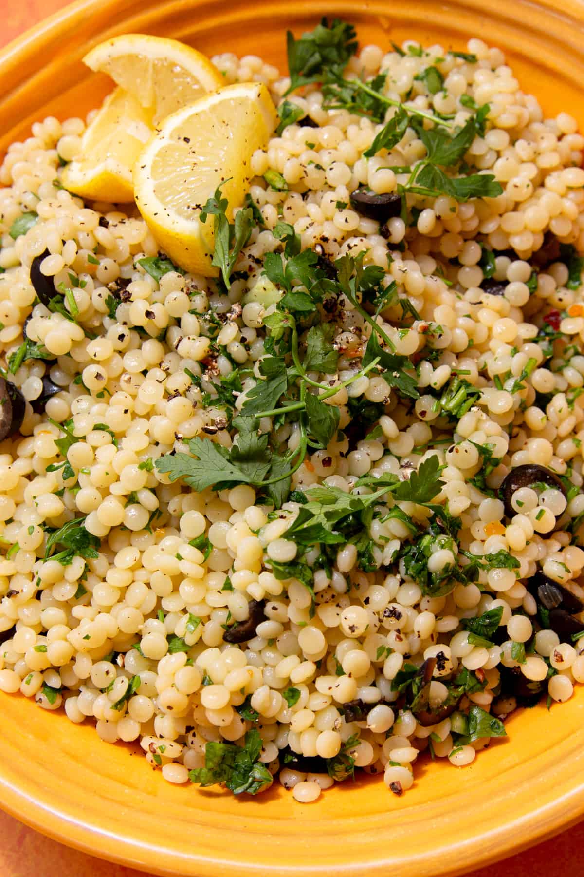 Close up shot of giant cous cous topped with herbs and lemon wedges and slices of black olives in a yellow bowl.
