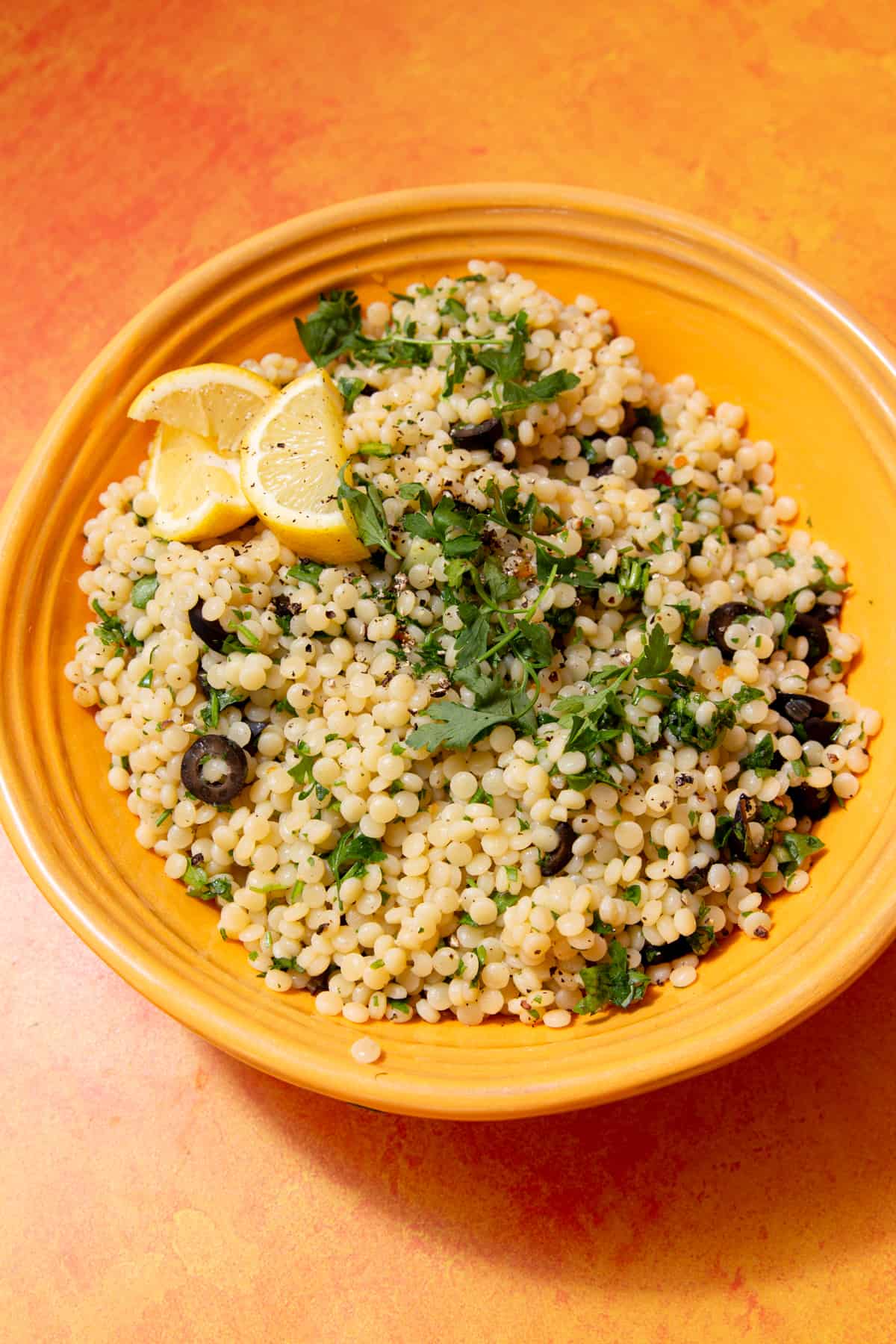 Overhead shot of a yellow dish with giant cous cous topped with herbs and lemon wedges and slices of black olives on an orange background.