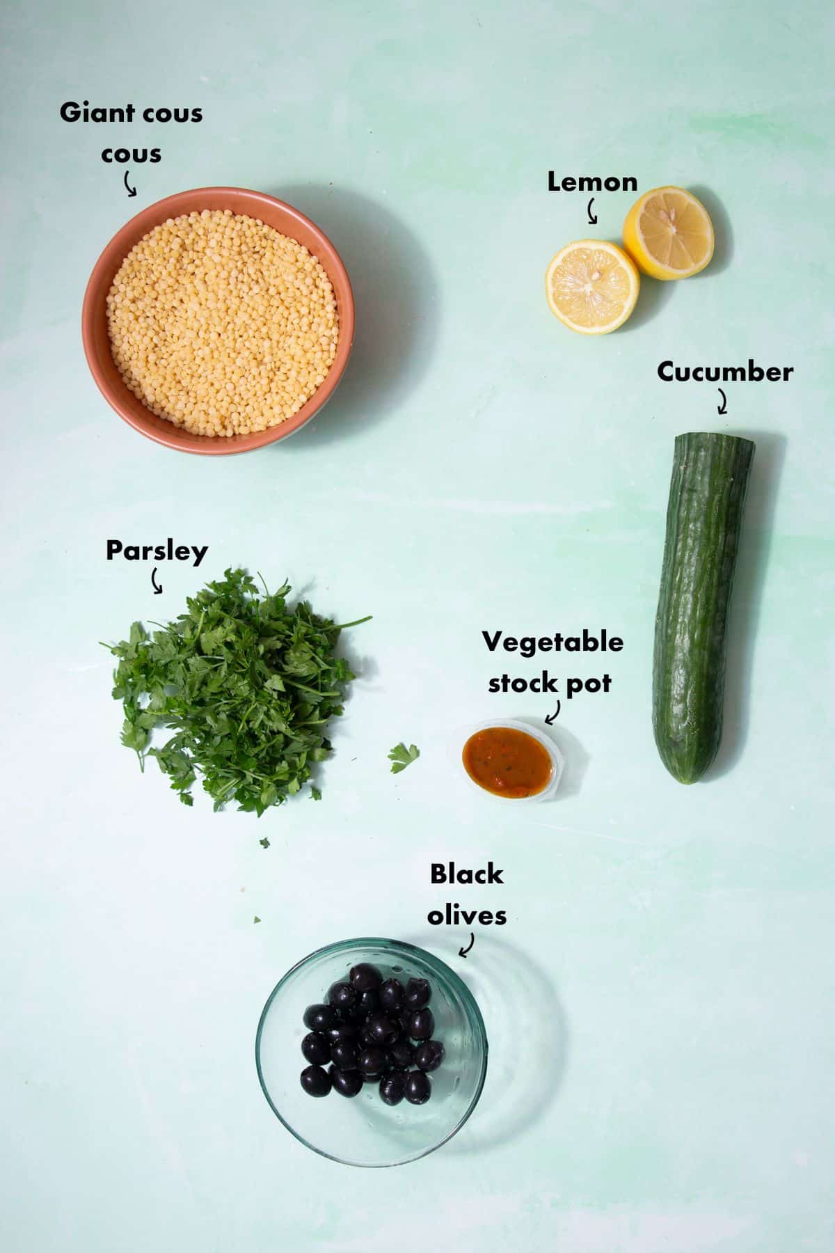 Ingredients to make the giant cous cous recipe laid out on a pale blue back ground and labelled.