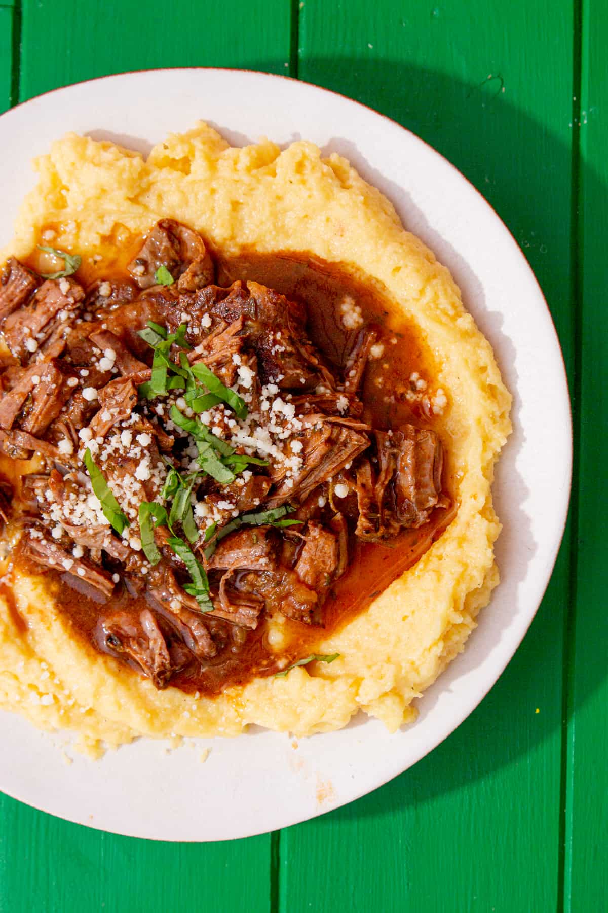 A plate with shredded steak with gravy over a bed of polenta on a green back ground.