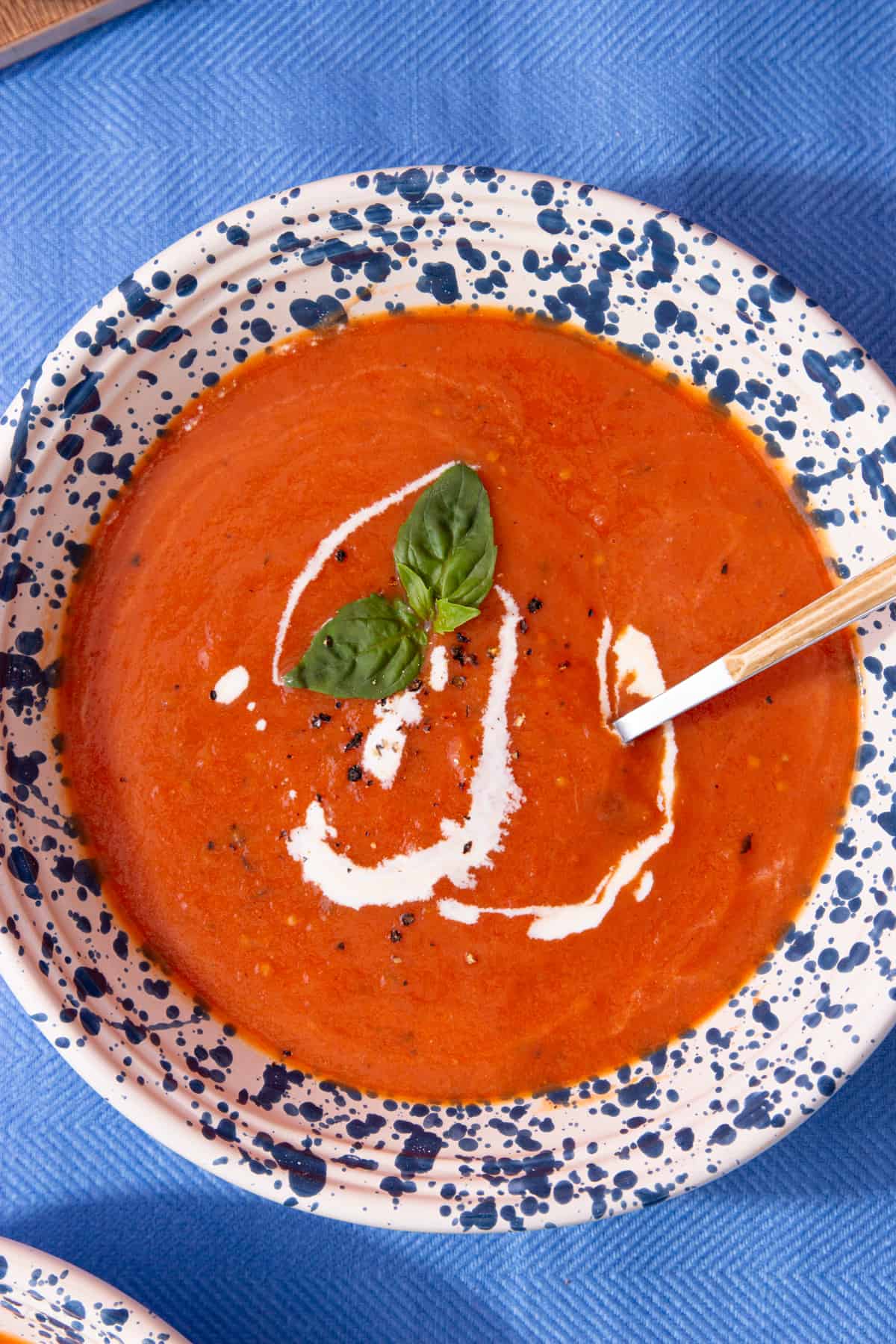 Overhead shot of tomato soup with a swirl of cream and some fresh basil in a cream bowl with blue spotty pattern on a blue cloth background.