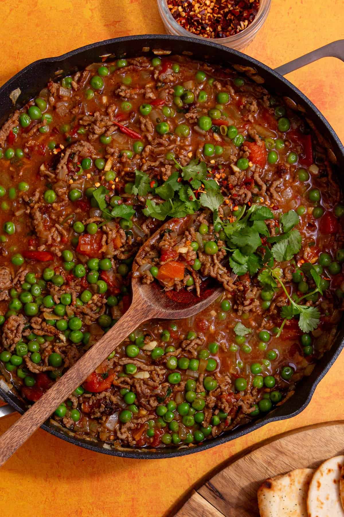 Mince curry with peas topped with fresh coriander with a wooden spoon in the pan and a small bowl of chillies behind the pan.