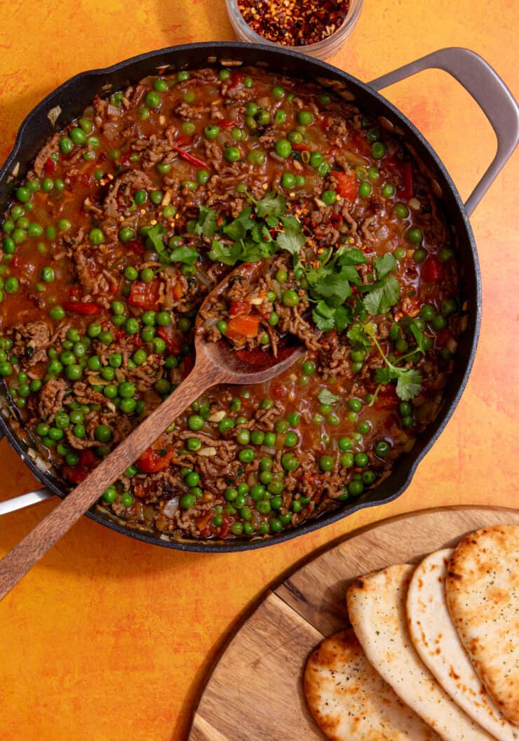 A large pan with mince curry with peas topped with coriander with a wooden spoon with naan breads.