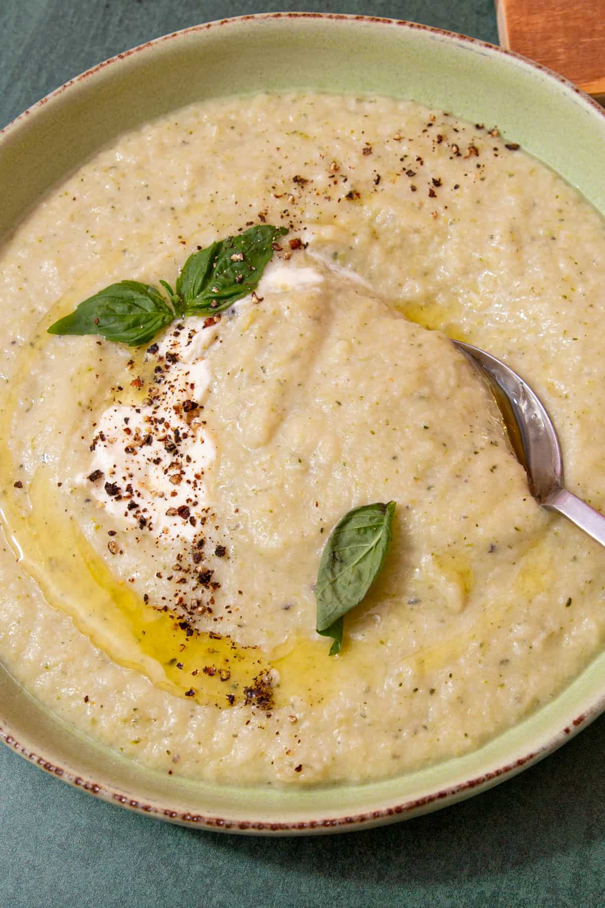 A bowl with a thick, creamy soup and a swirl of oil and cream topped with fresh basil and chilli flakes with a spoon.