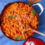Tomatoey, saucy fusilli pasta with chicken in a pan mixed with a wooden spoon and topped with fresh basil with a plate of parmesan.