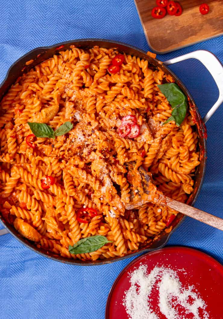 Tomatoey, saucy fusilli pasta with chicken in a pan mixed with a wooden spoon and topped with fresh basil with a plate of parmesan.
