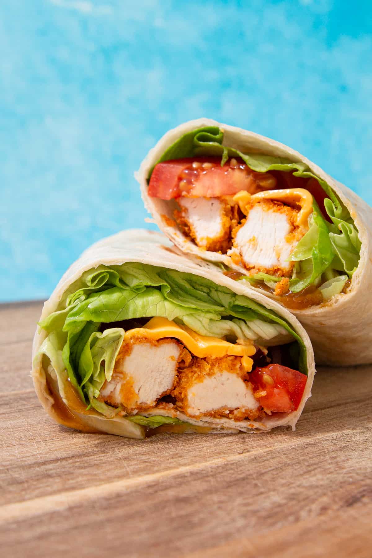 2 chicken wraps with lettuce, tomatoes, red onion piled one on top of the other over a wooden board on a blue background.