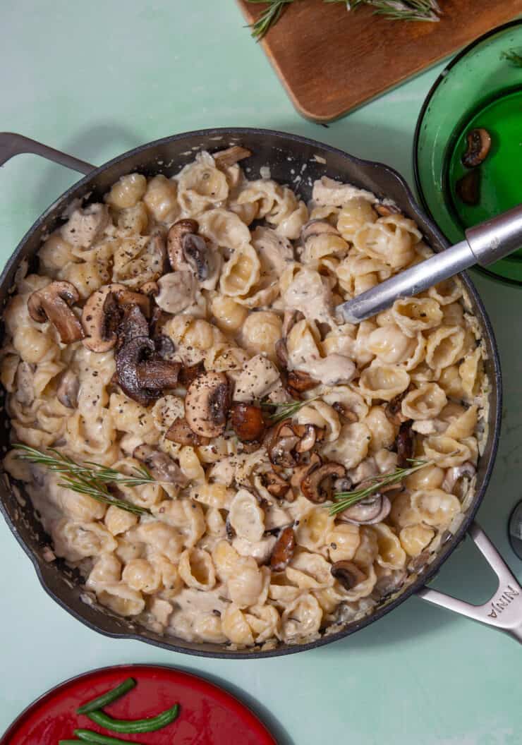 A large pan full of cream pasta shells topped with mushrooms and herbs with a metal spoon in the pan.