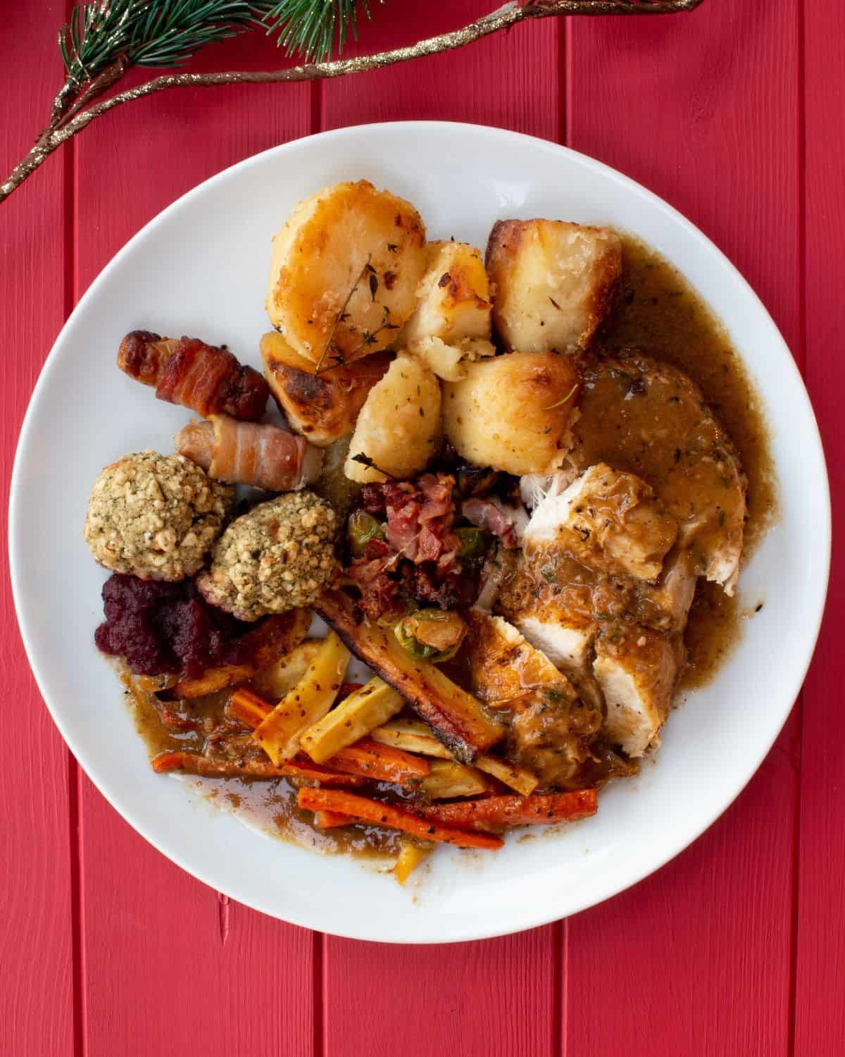 Christmas dinners with roasted potatoes, stuffing, pigs in blankets, roasted carrots and parsnips, chicken, brussel sprouts and cranberry sauce topped with gravy.