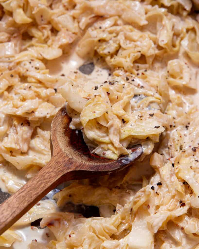 Creamy sliced cooked cabbage in a pan with a wooden spoon.