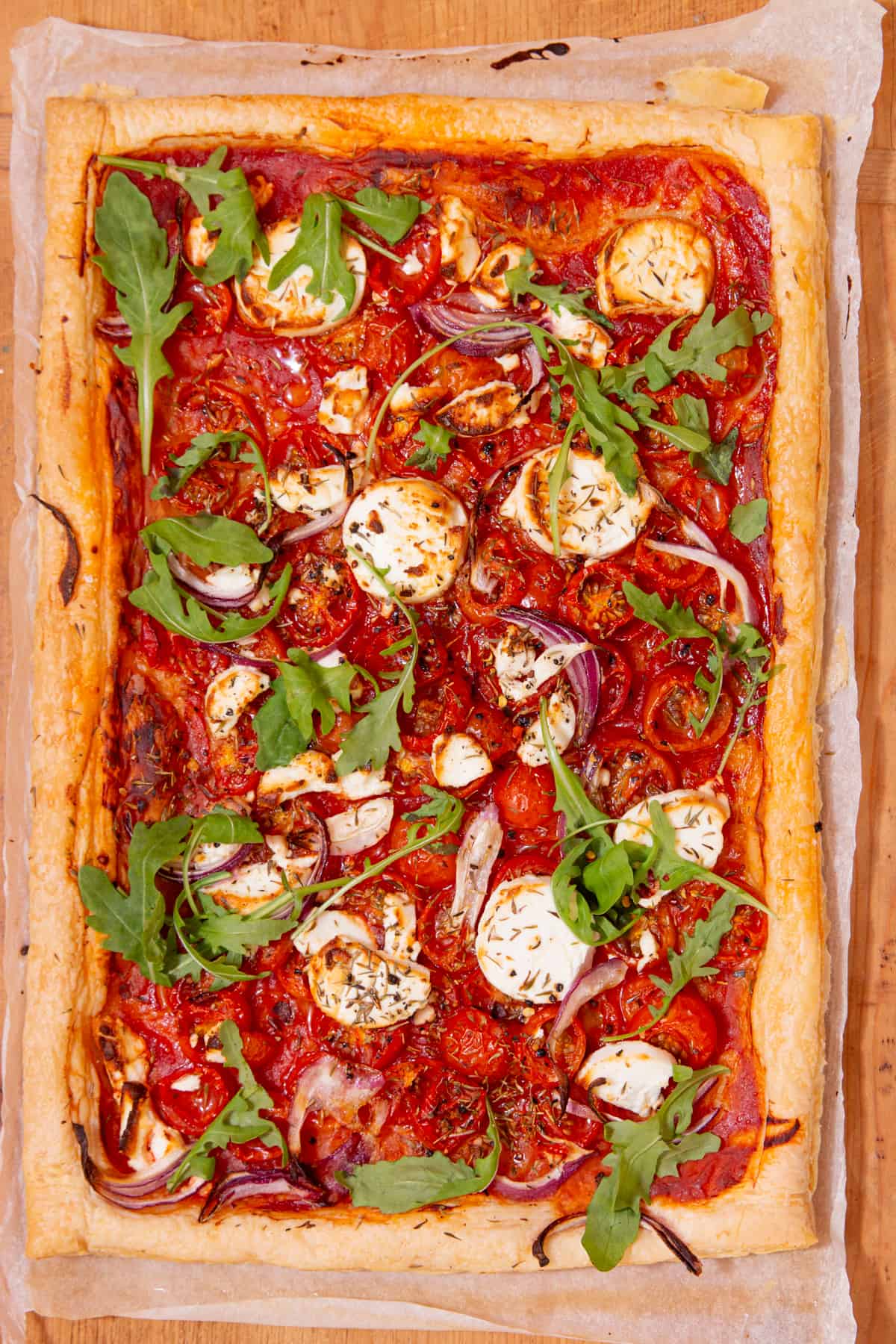 Rectangular, large goat's cheese tart with tomatoes and topped with rocket and red onion slices laid out on parchment paper on a wooden board.