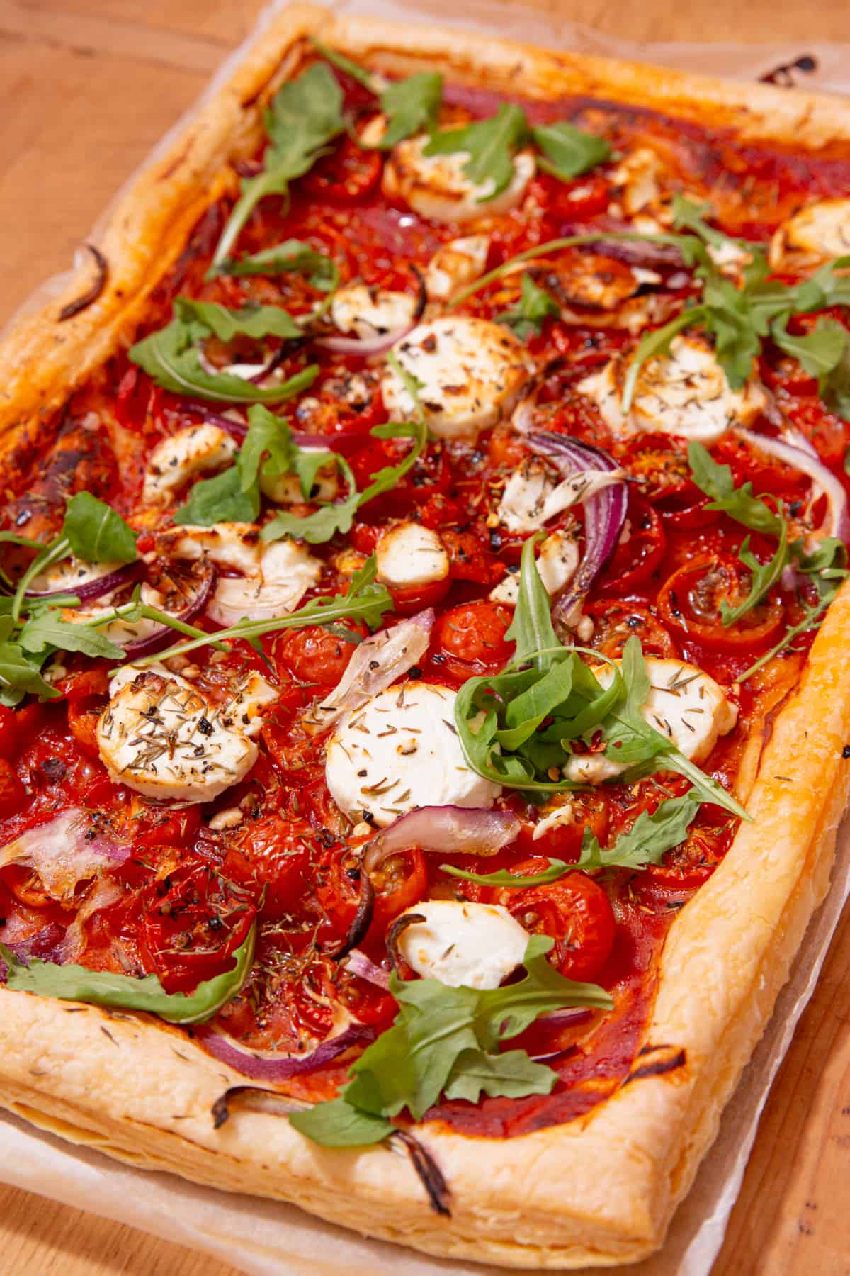 Large,rectangular pastry topped with goat's cheese  with tomatoes and topped with rocket and red onion slices laid out on parchment paper on a wooden board.