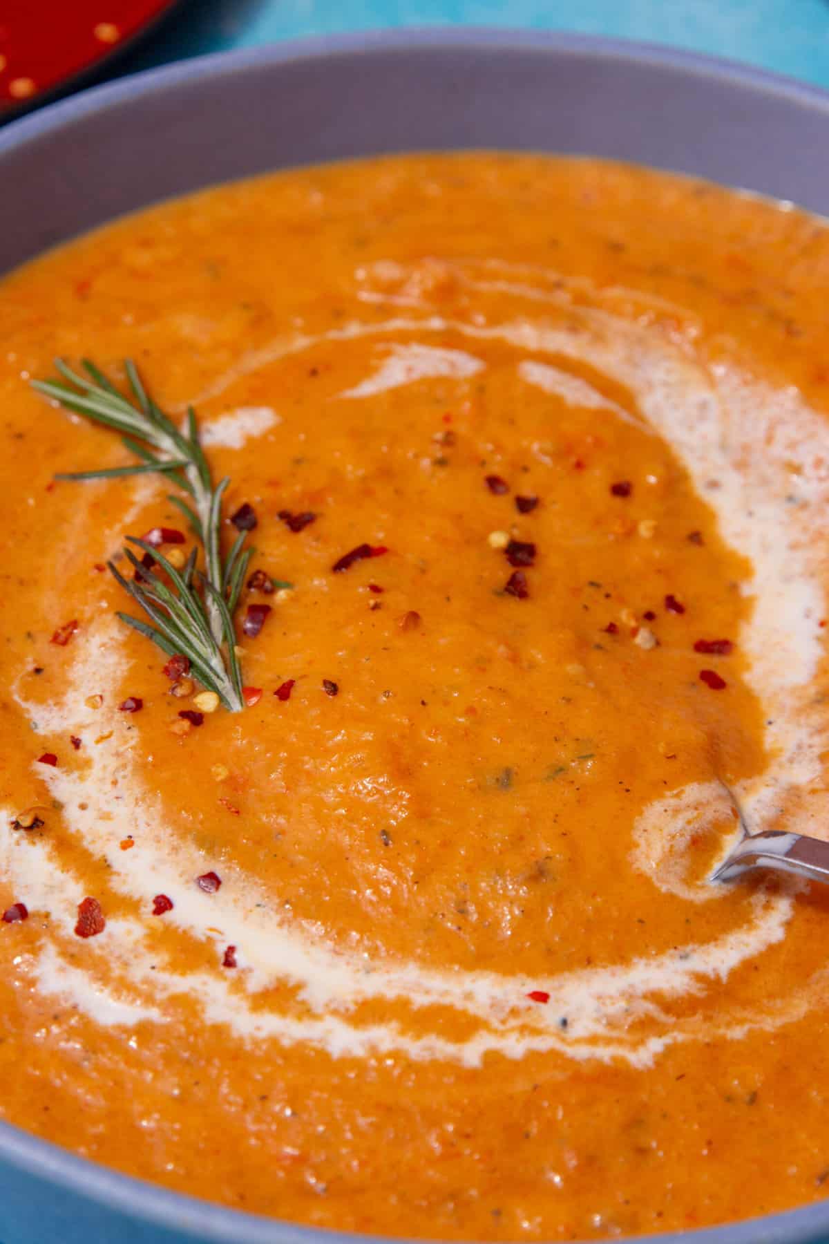 A blue bowl with an orangey thick vegetable soup with a swirl of cream, and topped with some chilli flakes and fresh rosemary.