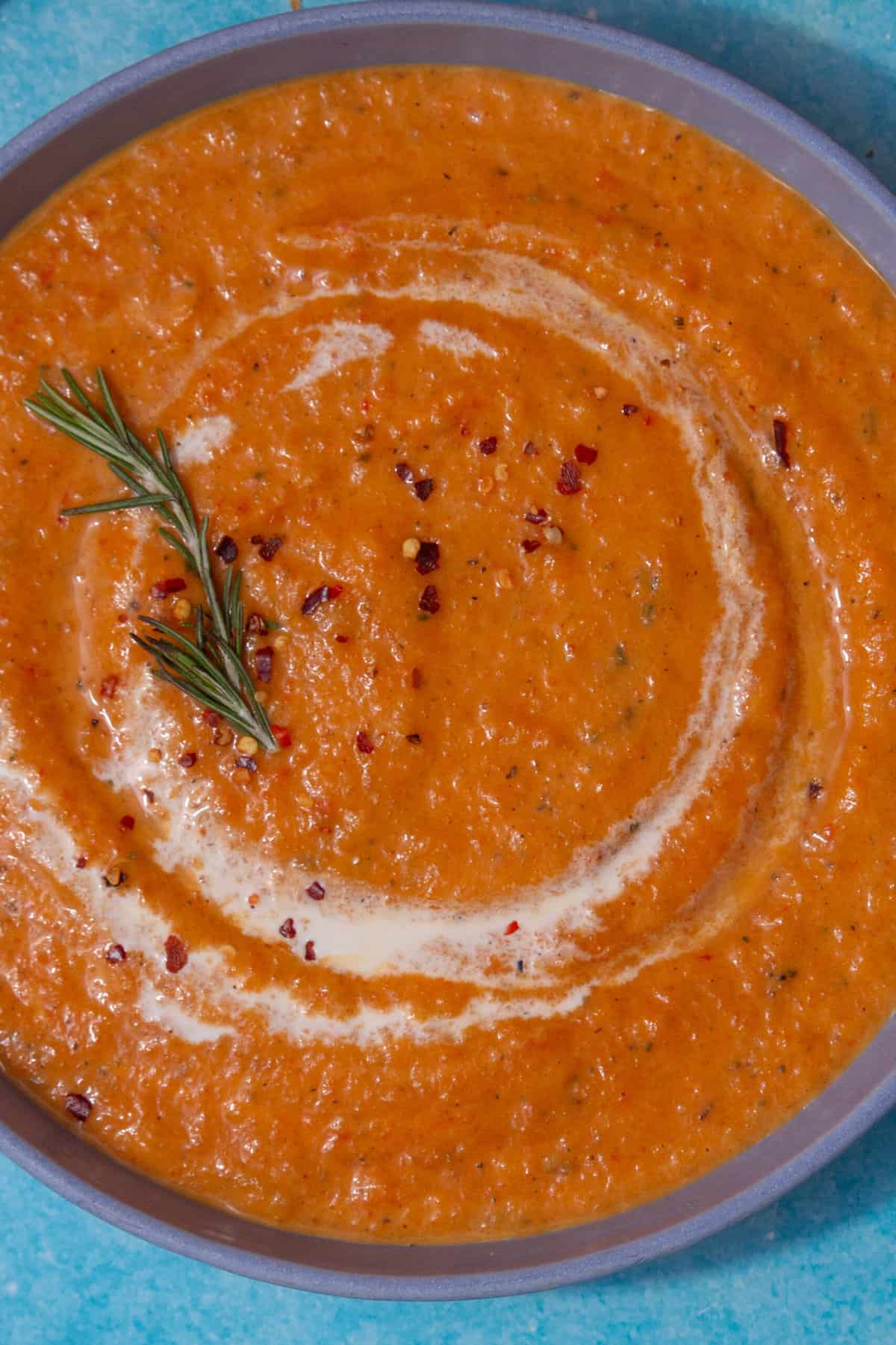 Orangey thick vegetable soup in a bowl with a swirl of cream, and topped with some chilli flakes and fresh rosemary.