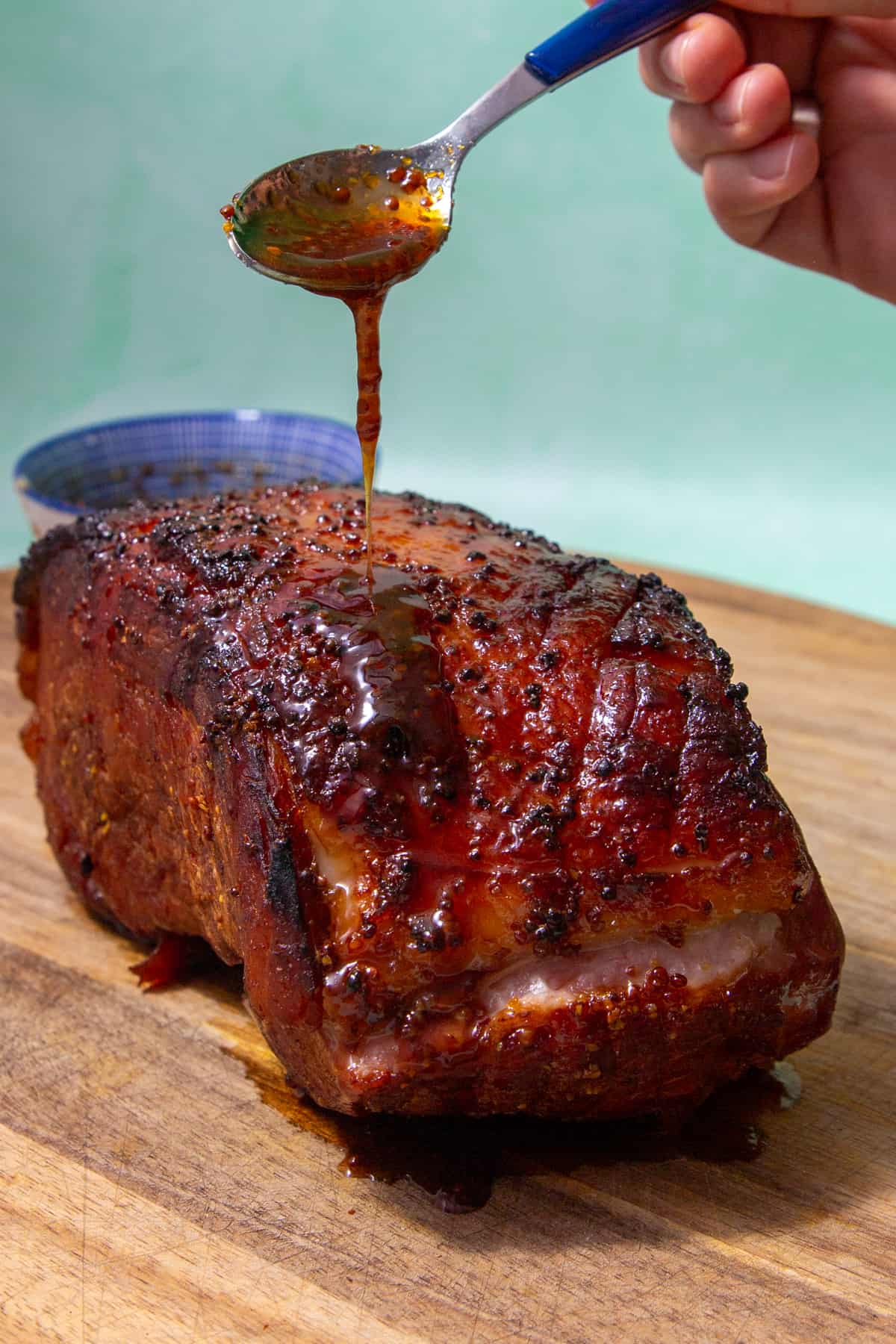 Roasted ham with a dark brown coating  witha spoon pouring the honey sauce over the top on a wooden board.