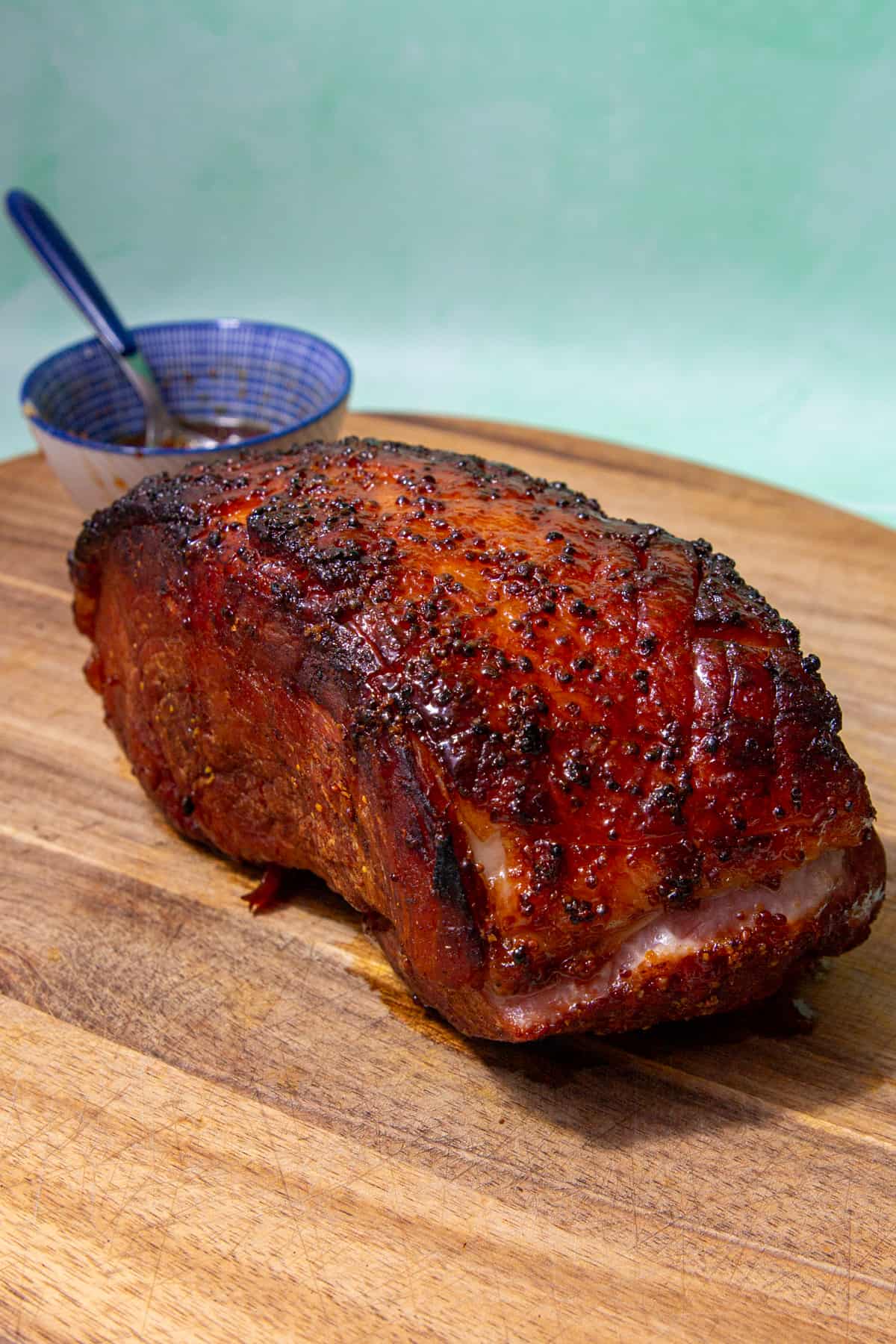 Roasted ham with dark brown glazed topping on a wooden board with a small pot in view behind the ham.