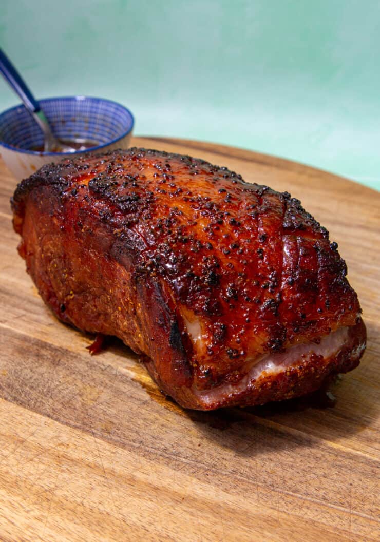 A joint of dark browned roasted/cooked gammon on a wooden board with a smal dish behind.