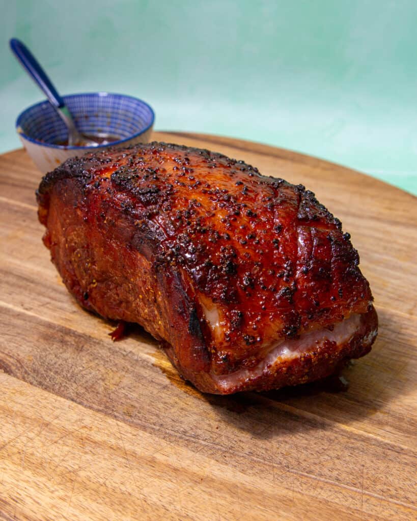 A joint of dark browned roasted/cooked gammon on a wooden board with a smal dish behind.