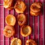 Golden brown Yorkshire puddings laid out on a metal grid on a red tile background.