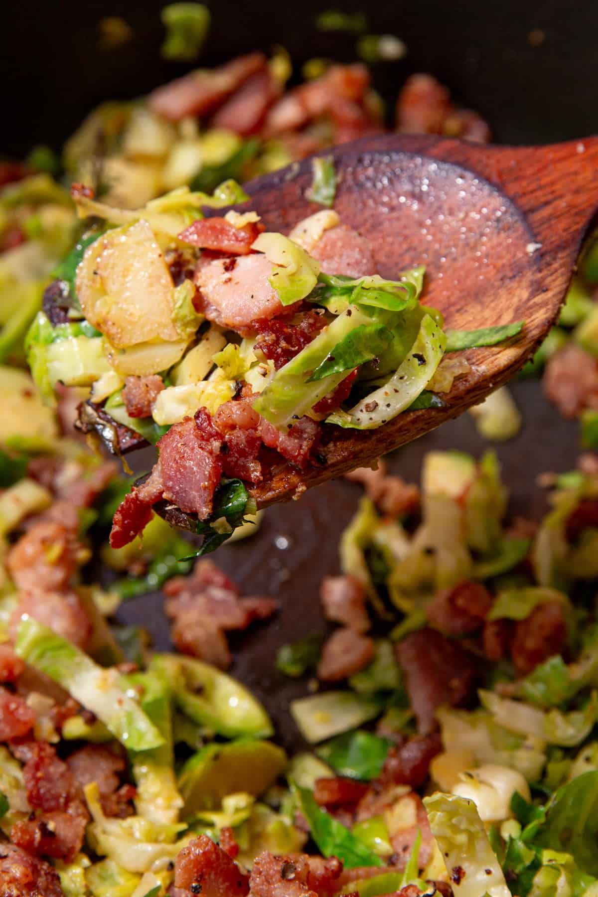 Sliced Brussel sprouts with browned bacon pieces being stirred with a wooden spoon.