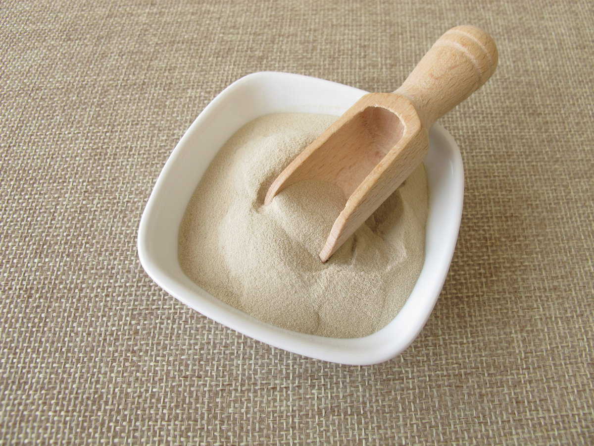 A beige grain in a square bowl with on wooden measuring spoon on a woven material background.