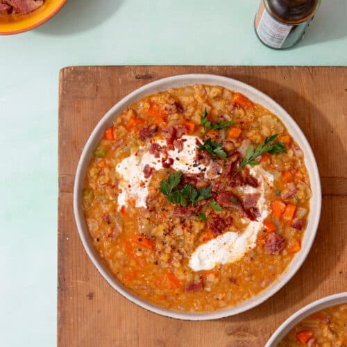 A bowl filled with lentil soup topped with bacon and sour cream on a wooden board with another 2 bowls in partial view.