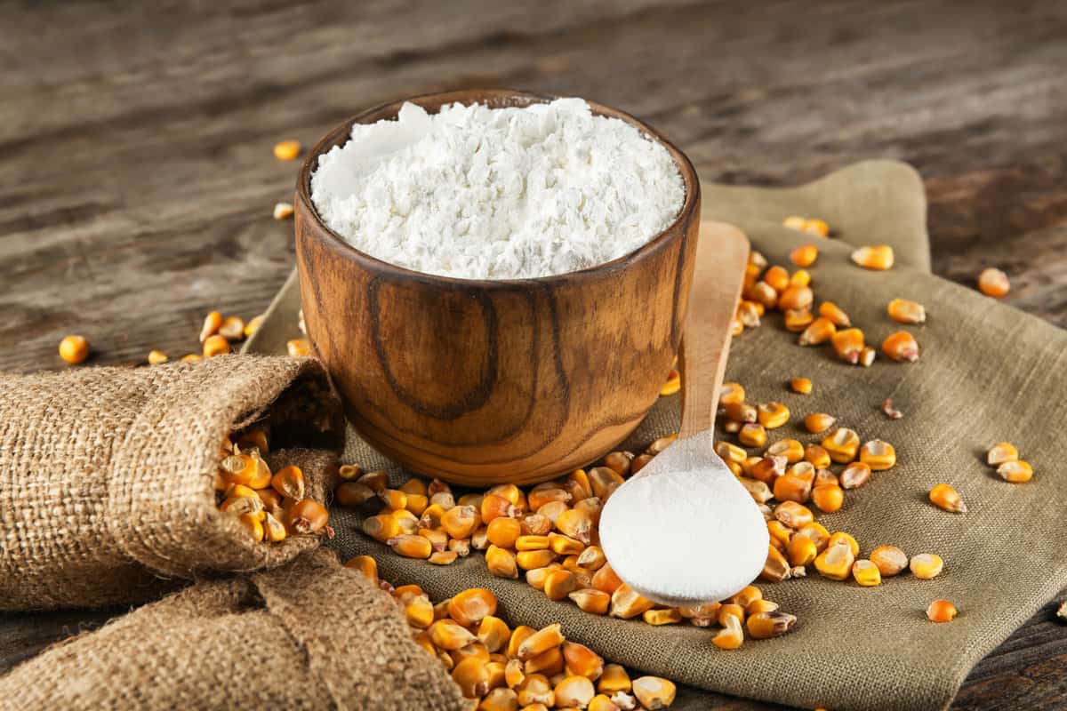 A deep wooden container with white cornstarch on a woven mat with corn kernels scattered over next to a wooden spoon.