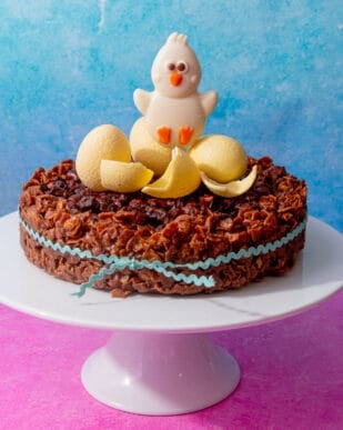 A Chocolate cornflake cake with white chocolate Easter eggs and one large chocolate bunny decorating the cake which is on a cake stand.