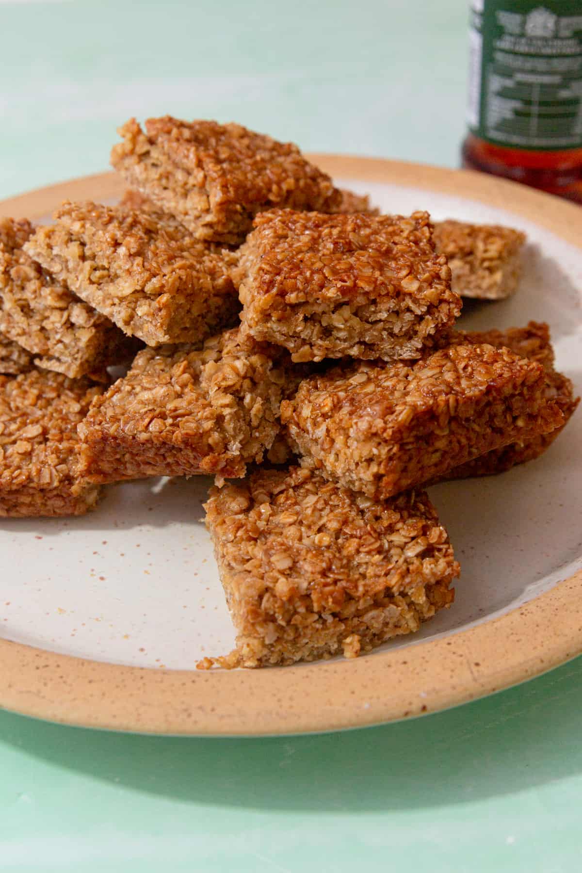 Lots of flapjack squares piled on a plate witha pale green background with a container of golden syrup in partial view.