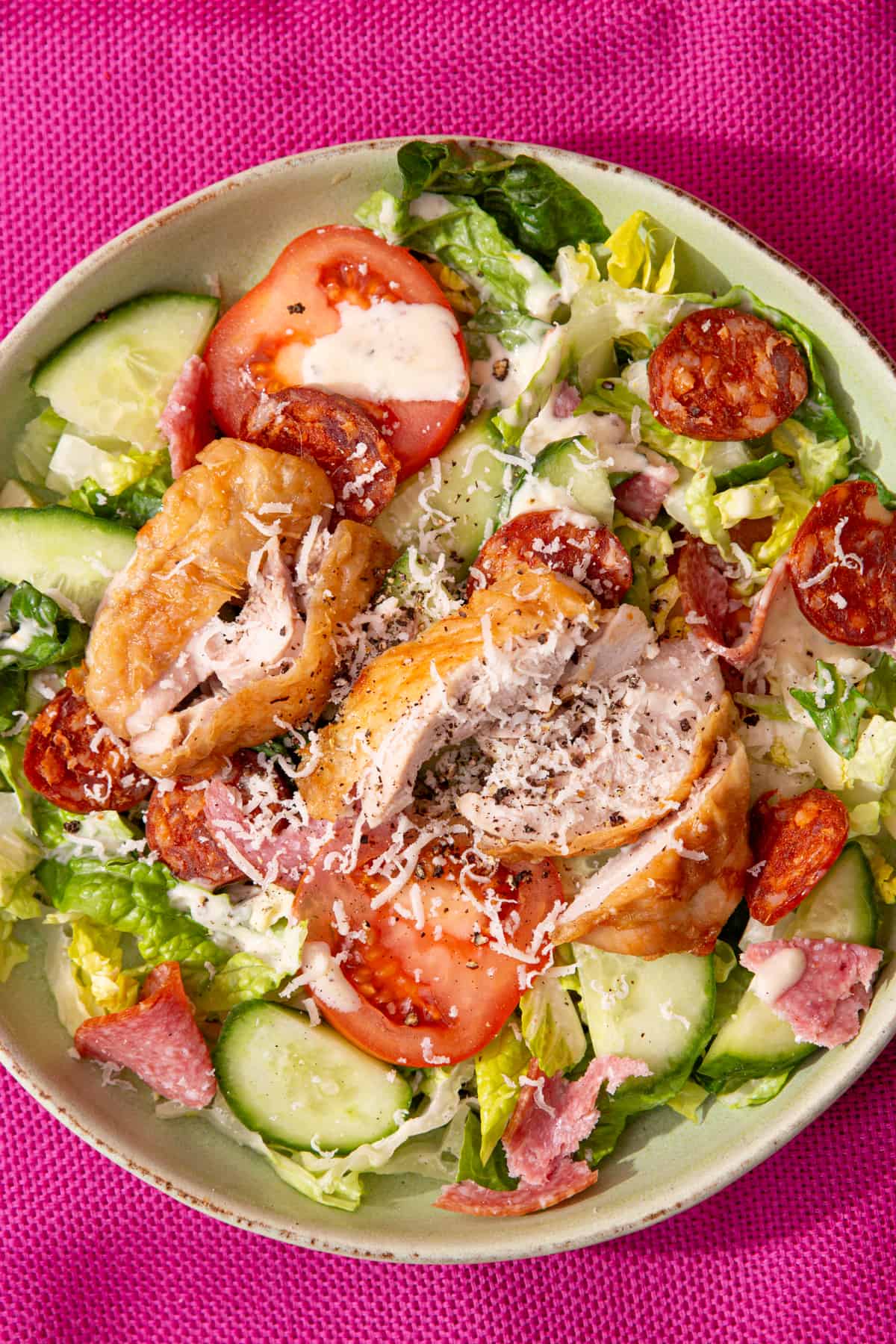 A bowl of salad with lettuce, tomatoes, cucumber, salami and chorizo pieces , chicken with skin slices and lots of parmesan shavings.