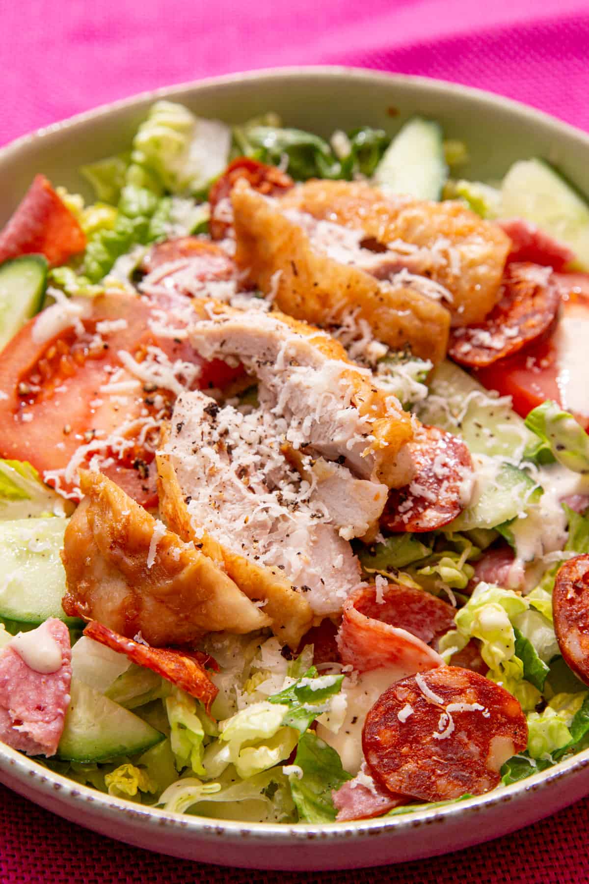 A bowl of salad with lettuce, tomatoes, cucumber, salami and chorizo pieces , chicken with skin slices and parmesan topping on a bright pink background. 