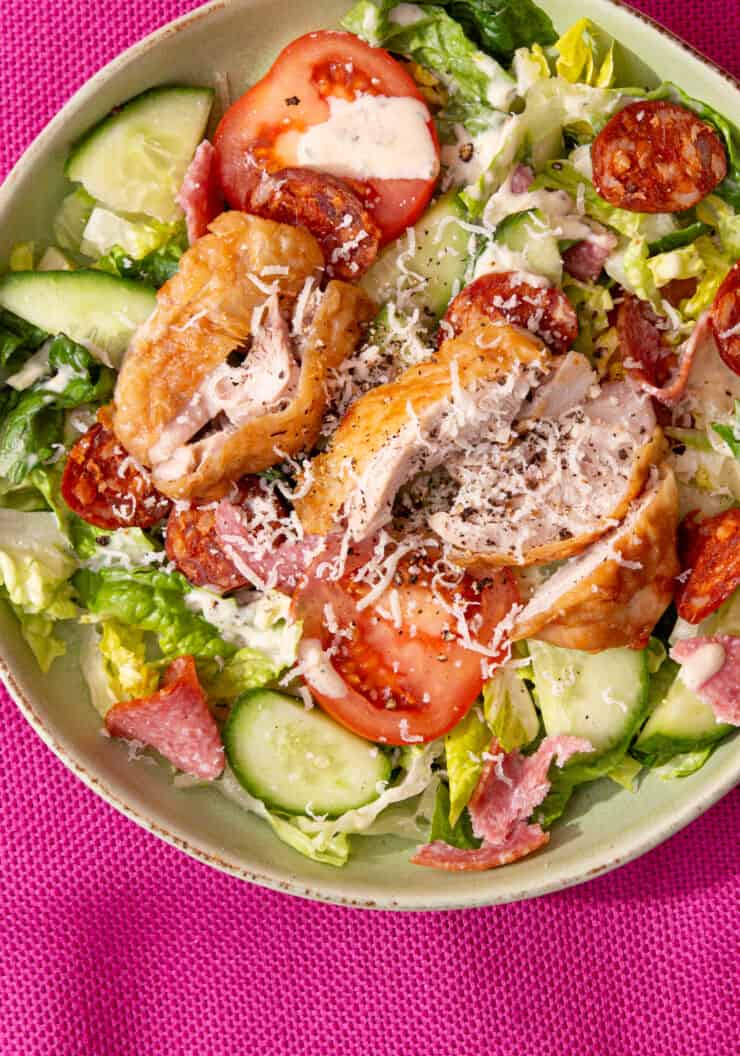 A bowl with salad, tomatoes, cucumber, meats, and a sprinkle of parmesan and dressing.