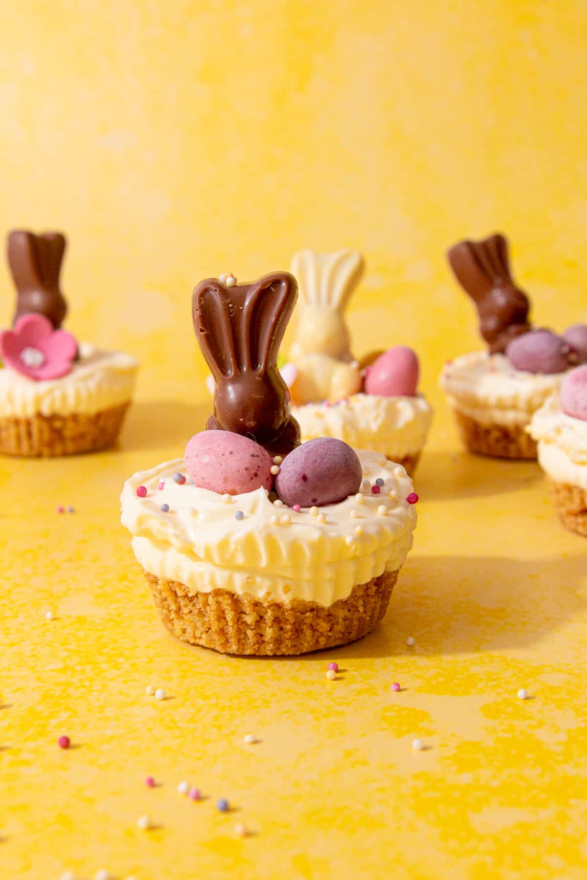 A few mini cheesecakes decorated with chocolate Easter bunnies, flowers and sprinkles on a yellow background.