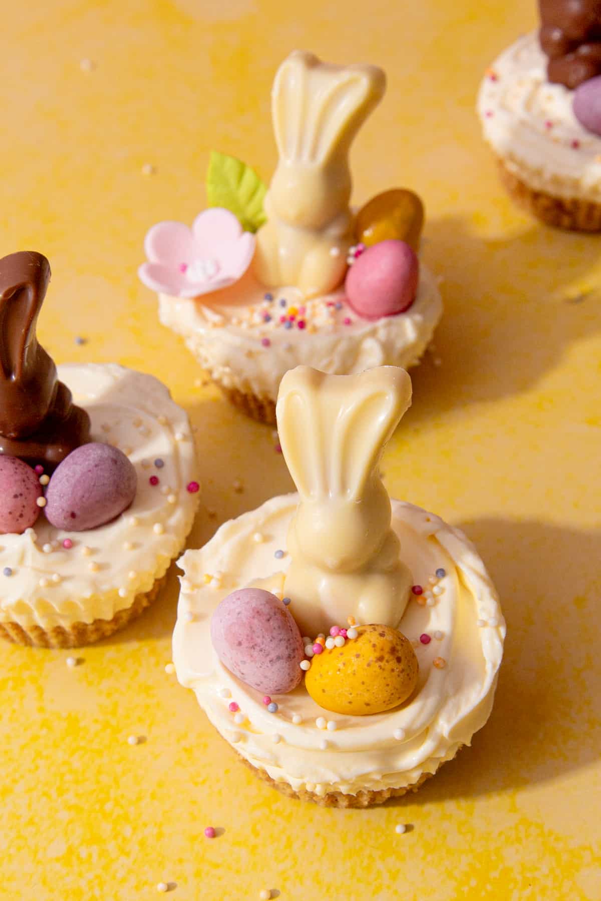Mini cheesecakes decorated with both milk and white chocolate Easter bunnies, flowers and sprinkles on a yellow background.