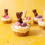 Mini cheesecakes topped with Easter chocolate bunnies, chocolate and sprinkles on a yellow background.