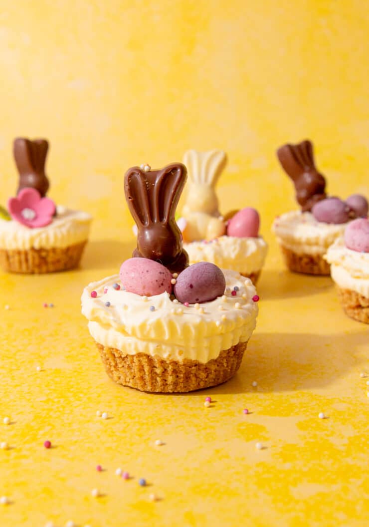 Mini cheesecakes topped with Easter chocolate bunnies, chocolate and sprinkles on a yellow background.