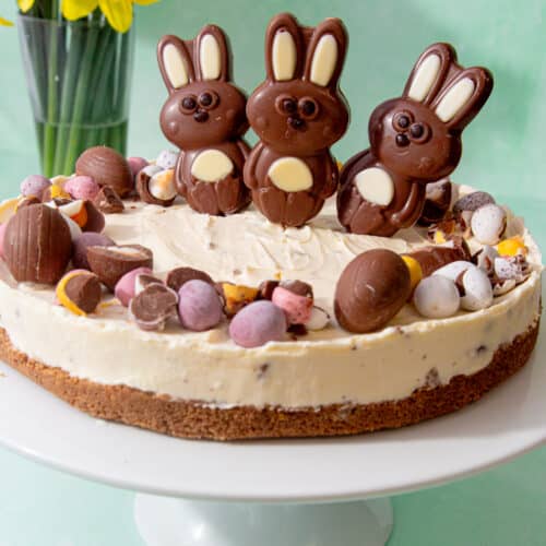 Cheese cake decorates with choclate mini eggs and 3 chocolate bunnies with some daffodils in the background.