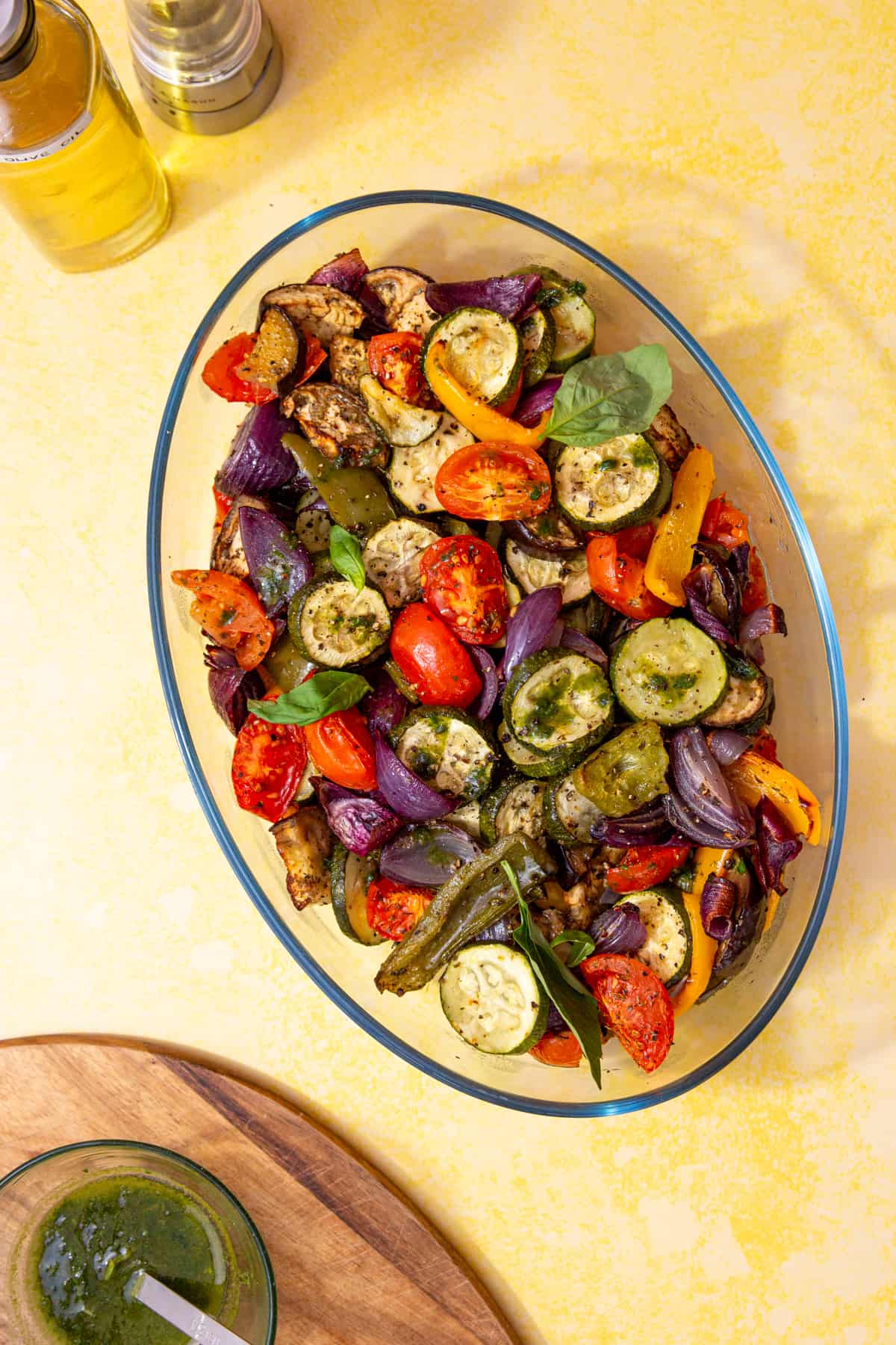 Roasted vegetables such a courgettes, tomatoes, peppers, red onions in a oval dish next to a bowl of green sauce mixture on a wooden board.