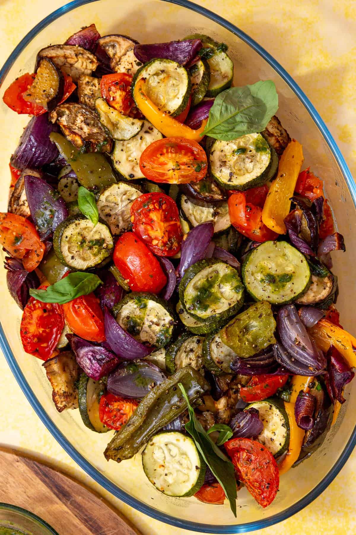 Roasted vegetables such a courgettes, tomatoes, peppers, red onions in a glass oval dish on a yellow background.